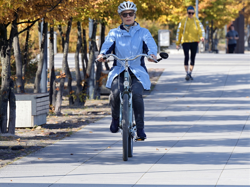 16 - A person wearing a white helmet and sunglasses while riding a light blue bicycle on a paved path surrounded by trees